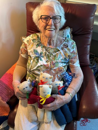 An elderly woman with glasses, sitting in a red chair, holding a collection of knitted dolls and smiling